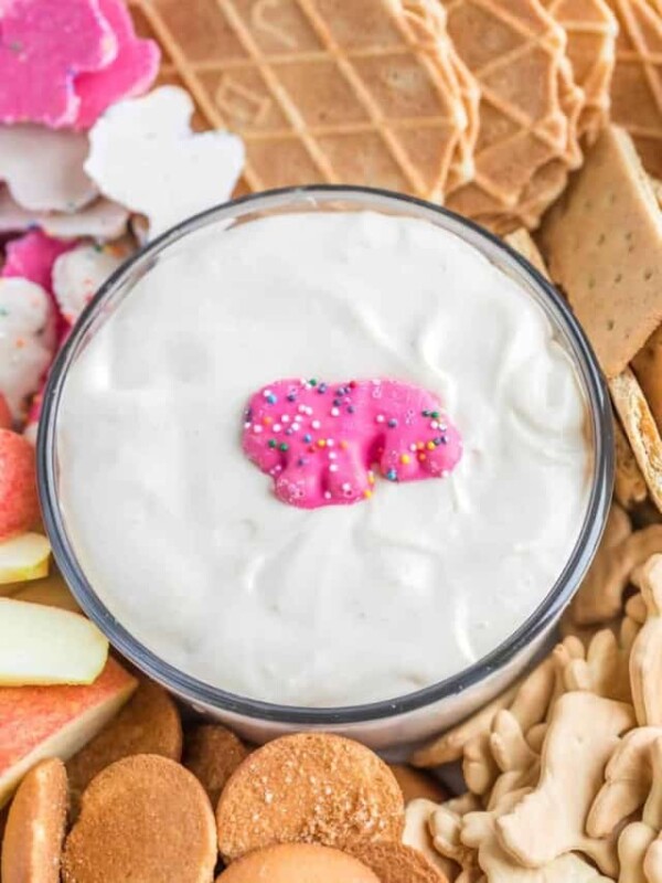 top view cropped close up of snacks and dip-able foods such as gram crackers, apples, and wafers with a silky white dip in the center wand an pink iced circus animal in the center