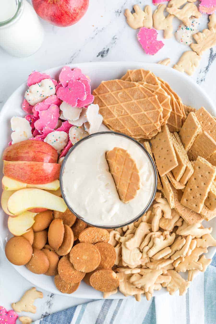 Top view of a bowl of cheesecake dip with a wafer in it on a plate surrounded by different cookies and apples.