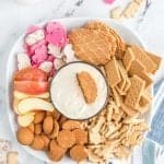 top view of a wafer dipped into the white cheesecake dip - top view of plated snacks and other dip-able foods such as gram crackers, apples, and wafers arranged around a glass bowl with a silky white cheesecake dip