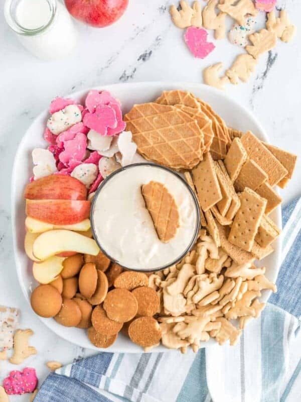 top view of a wafer dipped into the white cheesecake dip - top view of plated snacks and other dip-able foods such as gram crackers, apples, and wafers arranged around a glass bowl with a silky white cheesecake dip