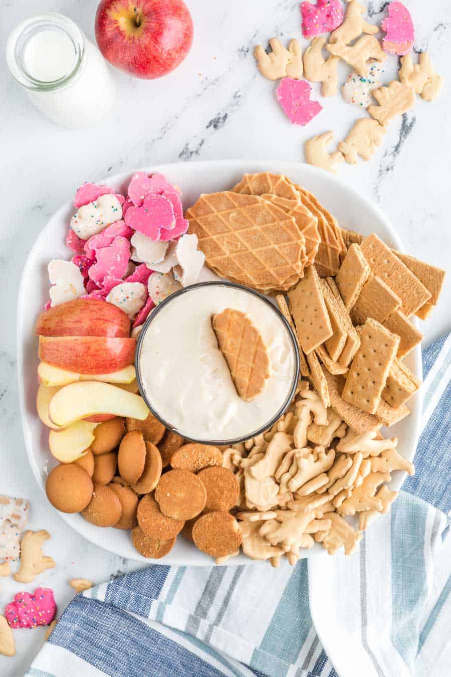 top view of a wafer dipped into the white cheesecake dip - top view of plated snacks and other dip-able foods such as gram crackers, apples, and wafers arranged around a glass bowl with a silky white cheesecake dip