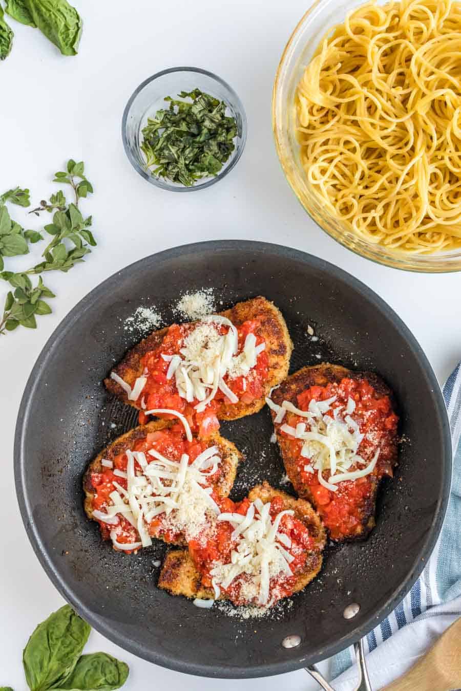 Top view of a skillet with chicken cooking, topped with a red sauce and white shredded cheese.