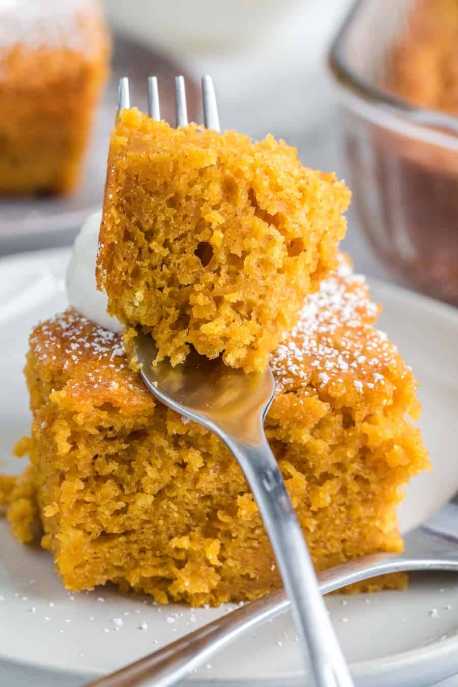 close up POV of a square serving of extra thick pumpkin cake on a white plate with a dollop of cream and one bite on a fork closest to the camera lens