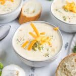 a marble table sets the scene for rustic white ceramic bowls of broccoli cheddar soup with rolls and raw broccoli around