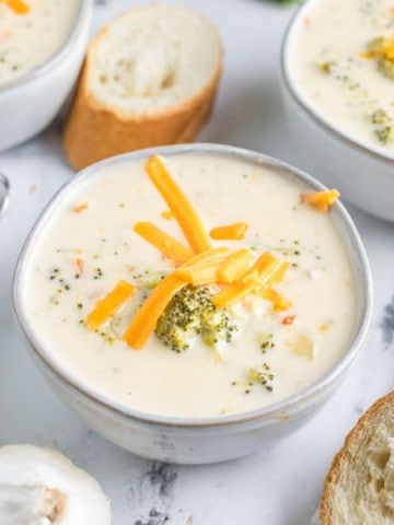a marble table sets the scene for rustic white ceramic bowls of broccoli cheddar soup with rolls and raw broccoli around