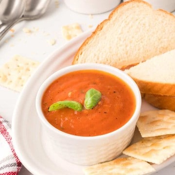 roasted tomato soup in a white cup on a white plate with a sandwich crackers, spoon, and a sprig of basil