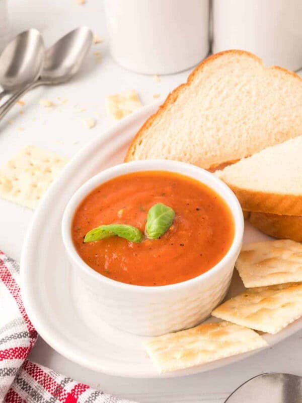 roasted tomato soup in a white cup on a white plate with a sandwich crackers, spoon, and a sprig of basil