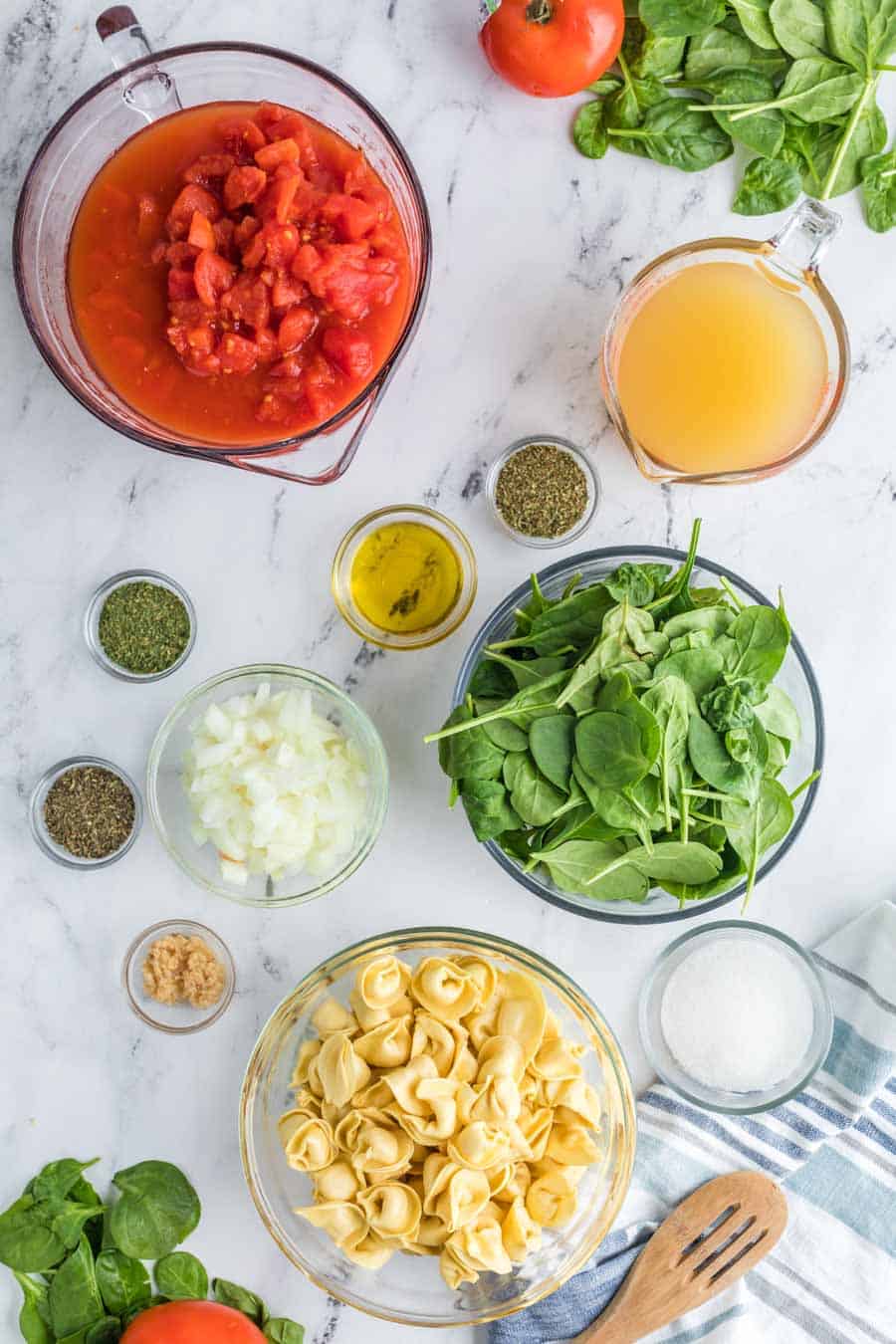 glass bowls and measuring dishes over marble table aesthetically arranged - ingredients like tortellin - stock - tomatoes - spinach - and spices