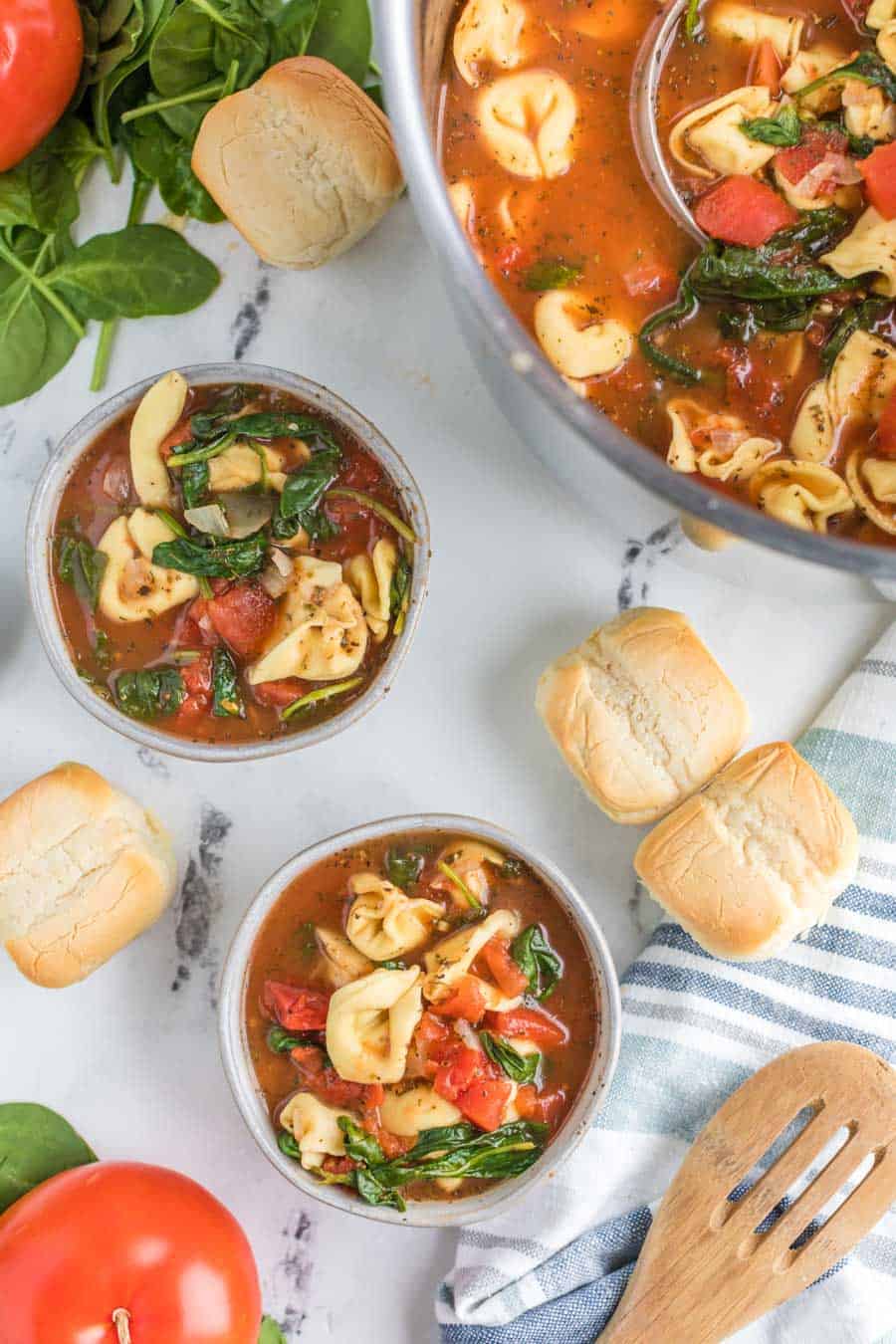 completed bowls of tortellini tomato soup in a scene over marble table with rolls, whole tomatoes, a wooden spoon, and the edge of the large pot of soup with a ladle in it