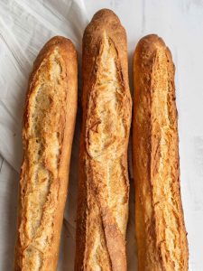 Three golden, crusty baguettes—made from a classic baguette recipe—are placed side by side on a white surface, with a white cloth partially visible in the background.