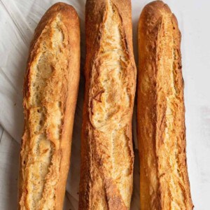 Three golden-brown baguettes, made from a classic baguette recipe, with crisp, textured crusts rest side by side on a white surface, partially overlapping a white cloth.