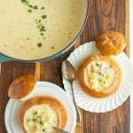 top view of a creamy chowder soup and two plates next to it with crunchy round bread bowls on white plates with the top perfectly cut off and chunky cream soup and a spoon