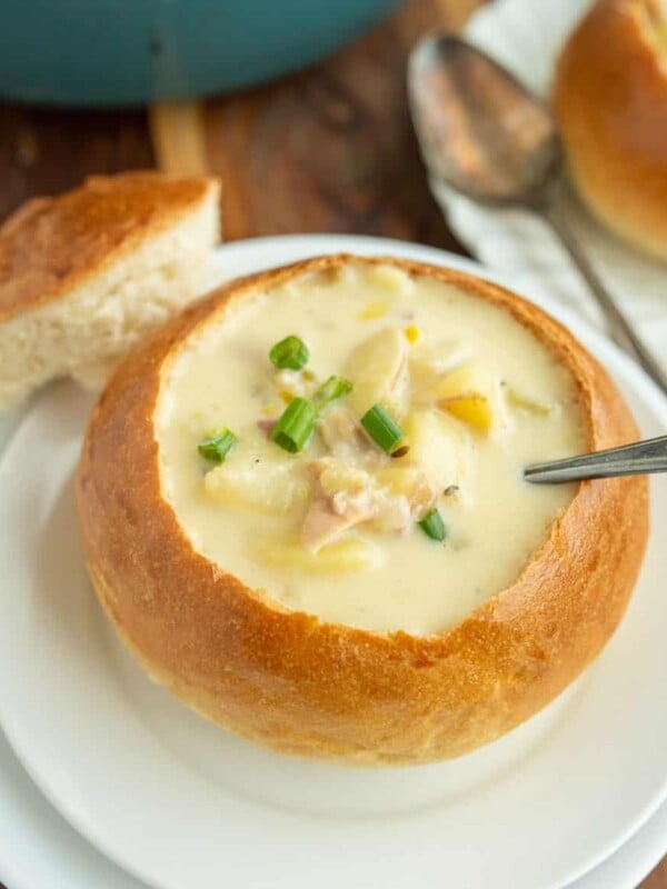 white plate holding a large crunchy round bread soup bowl with chunky chowder the center and a silver spoon