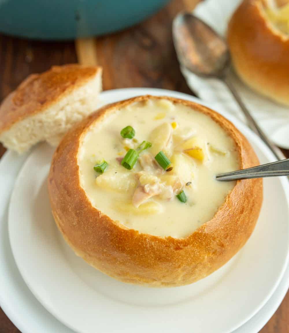 white plate holding a large crunchy round bread soup bowl with chunky chowder the center and a silver spoon.