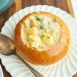 wooden board and a shell pattern white plate holding a large crunchy round bread soup bowl with chunky cream souping the center and a silver spoon to the left