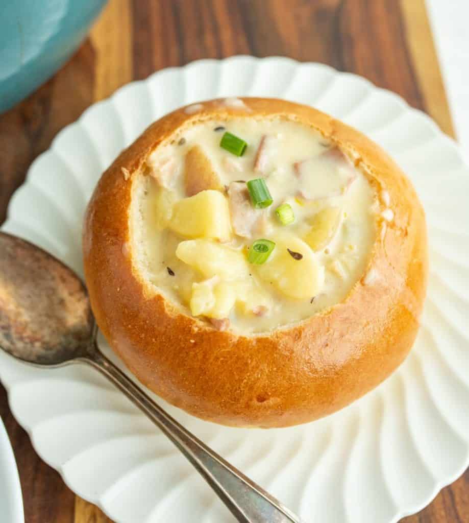 wooden board and a shell pattern white plate holding a large crunchy round bread soup bowl with chunky cream souping the center and a silver spoon to the left