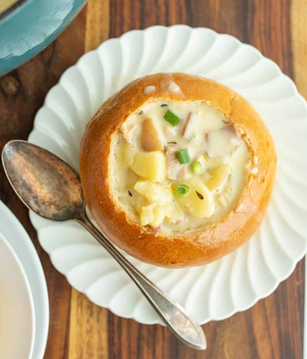 wooden board and a shell pattern white plate holding a large crunchy round bread soup bowl with chunky cream souping the center and a silver spoon to the left.