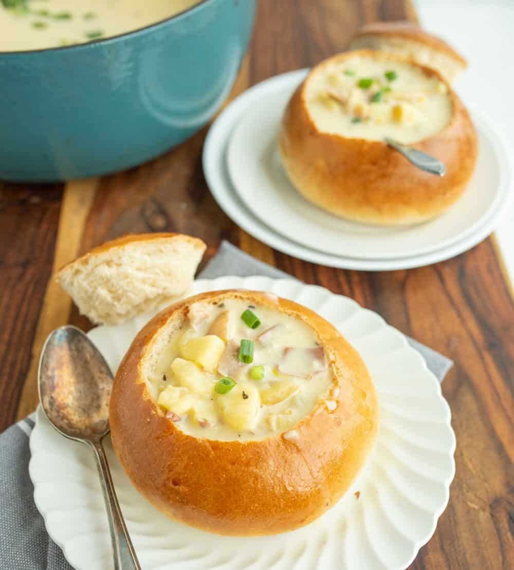 crunchy round bread bowl on white plate with the top perfectly cut off and chunky cream soup and a spoon in it a second plate in the background and a rustic turquoise soup pot.