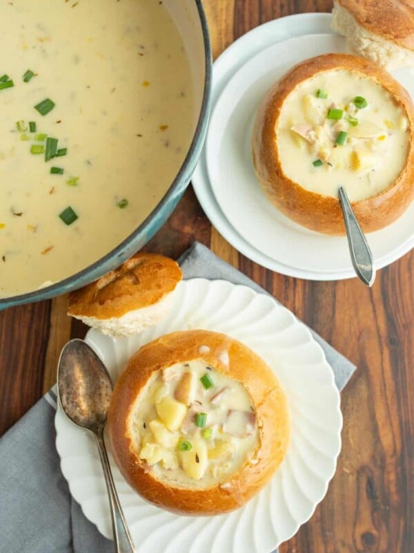 top view of a creamy chowder soup and two plates next to it with crunchy round bread bowls on white plates with the top perfectly cut off and chunky cream soup and a spoon