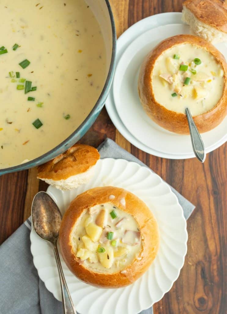 top view of a creamy clam chowder with canned clams soup and two plates next to it with crunchy round bread bowls on white plates with the top perfectly cut off and chunky cream soup and a spoon.