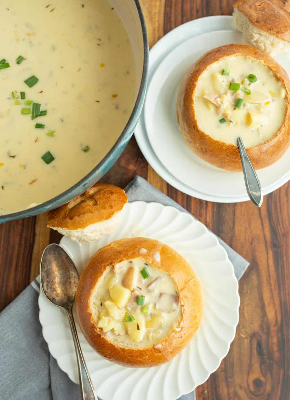 top view of a creamy chowder soup and two plates next to it with crunchy round bread bowls on white plates with the top perfectly cut off and chunky cream soup and a spoon.