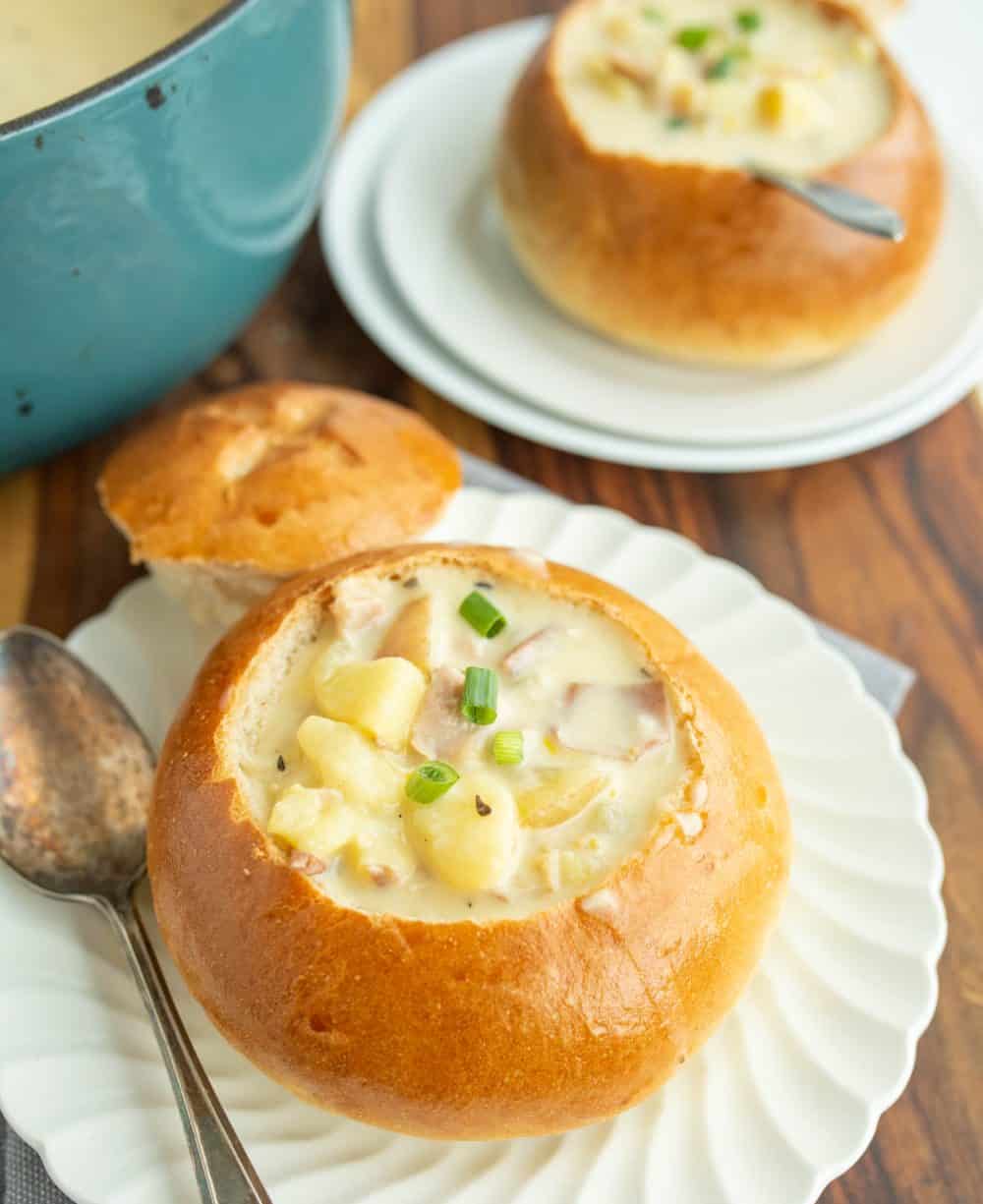 crunchy round bread bowl on white plate with the top perfectly cut off and chunky cream soup and a spoon in it a second plate in the background