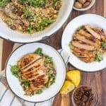 wide angle of the wooden block, the beautiful quinoa salad in a bowl, and two completed dishes - perfectly cooked chicken breast with a seared top sliced horizontal laid atop a quinoa salad with whole spinach leaves in a rustic white ceramic dish