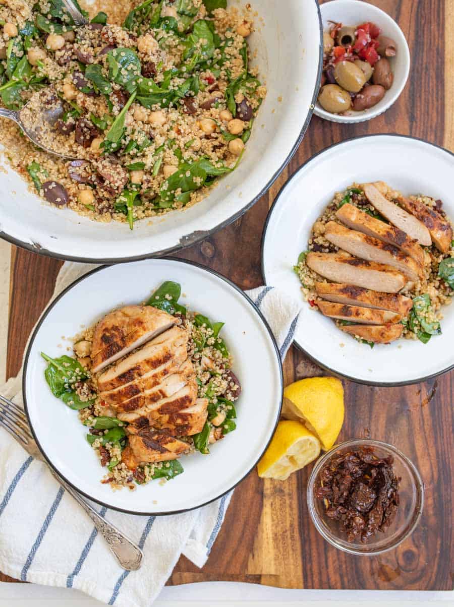 wide angle of the wooden block, the beautiful quinoa salad in a bowl, and two completed dishes - perfectly cooked chicken breast with a seared top sliced horizontal laid atop a quinoa salad with whole spinach leaves in a rustic white ceramic dish