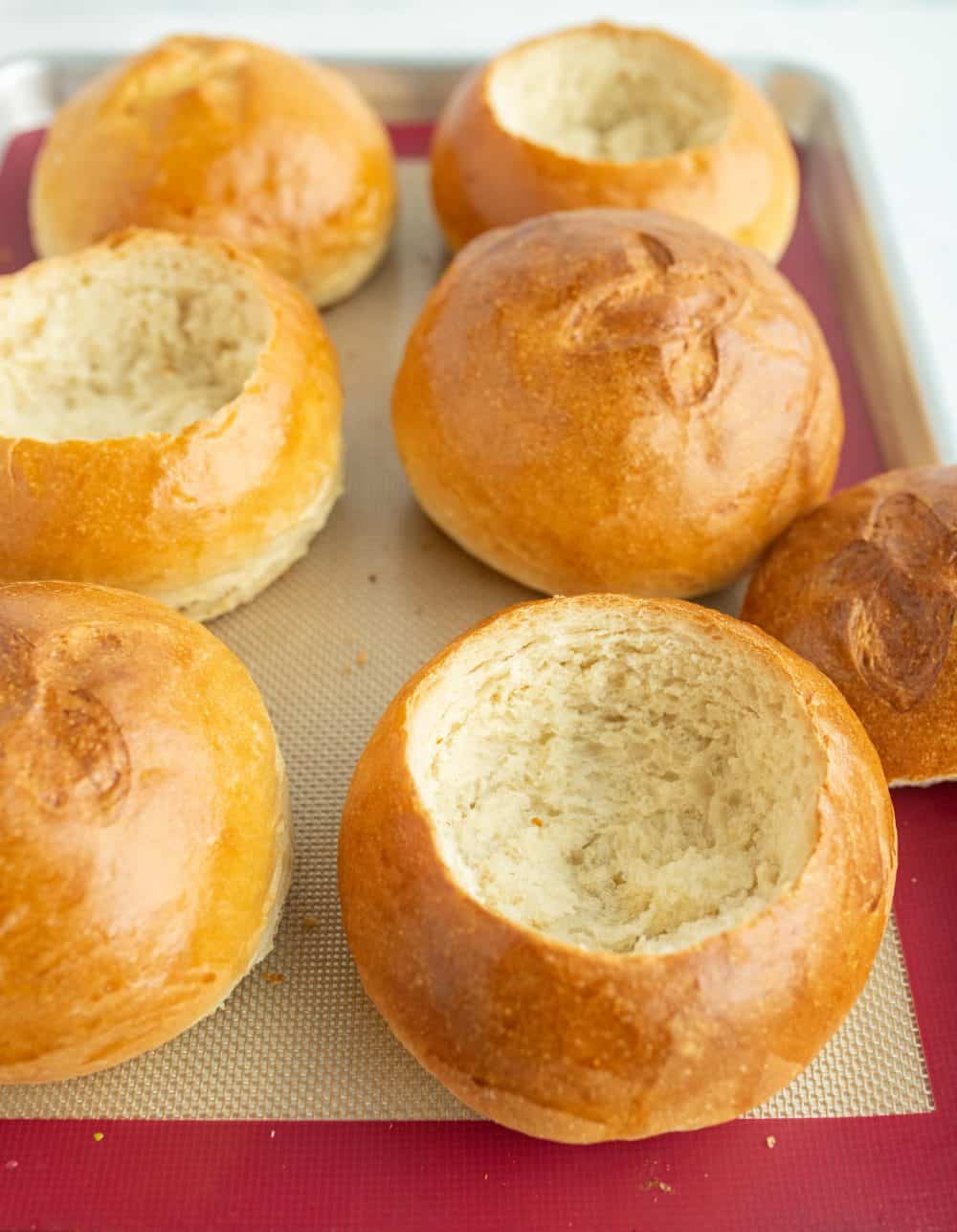 close up 6 large round rolls baked golden brown and a little x on the tops line a silicone baking sheet and a few of the rolls are hollowed for the bread bowl.