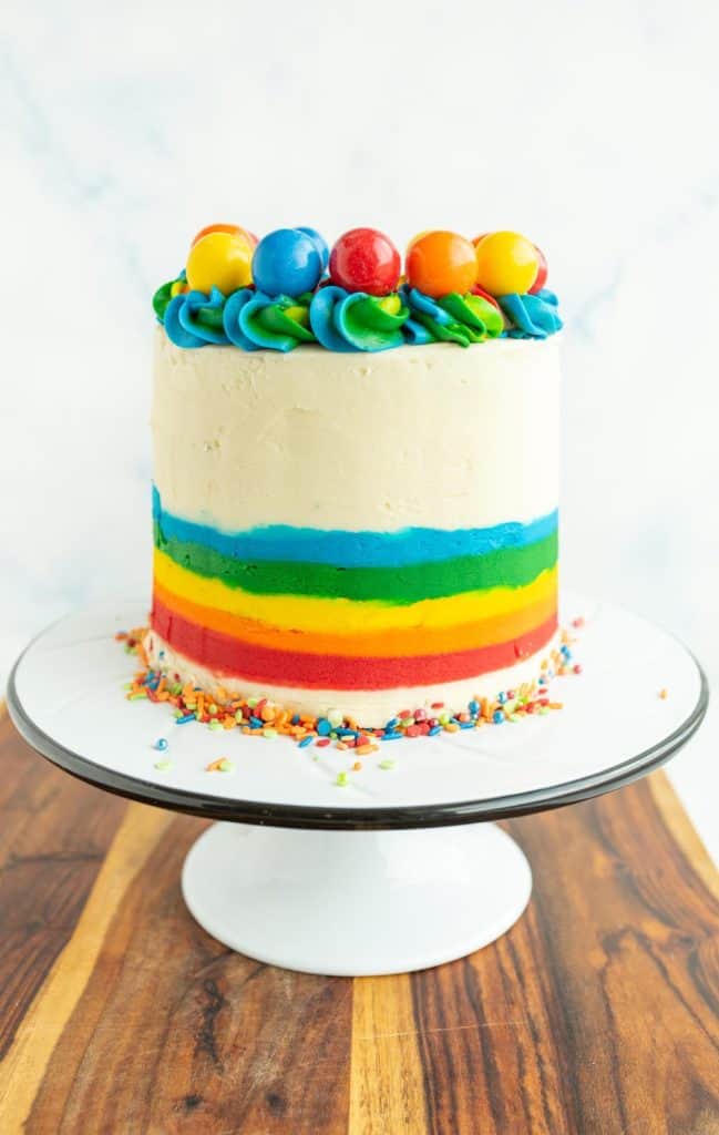 wooden board with a white stand and the rainbow cake is whole and the outer icing is decorated with the bottom half as a rainbow and top ¾ white icing and along the top of the cake is rainbow piped dollops with different colorful gumboils on top