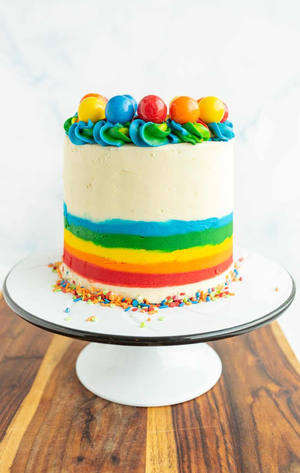 wooden board with a white stand and the rainbow cake is whole and the outer icing is decorated with the bottom half as a rainbow and top 3/4 white icing and along the top of the cake is rainbow piped dollops with different colorful gumboils on top