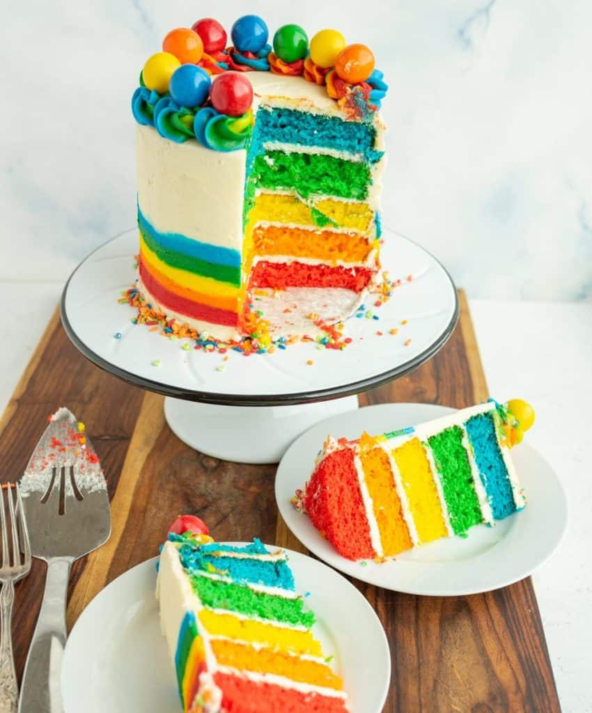 vibrant rainbow layered cake with elaborate piping staged over a wooden board on white dishes - the whole cake is on a stand to the back and it has two slices cut out that are on plates in the foreground