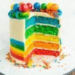 close up of the round rainbow layer cake on a white stand - the outer icing is decorated with the bottom half as a rainbow and top 3/4 white icing and along the top of the cake is rainbow piped dollops with different colorful gumboils on top