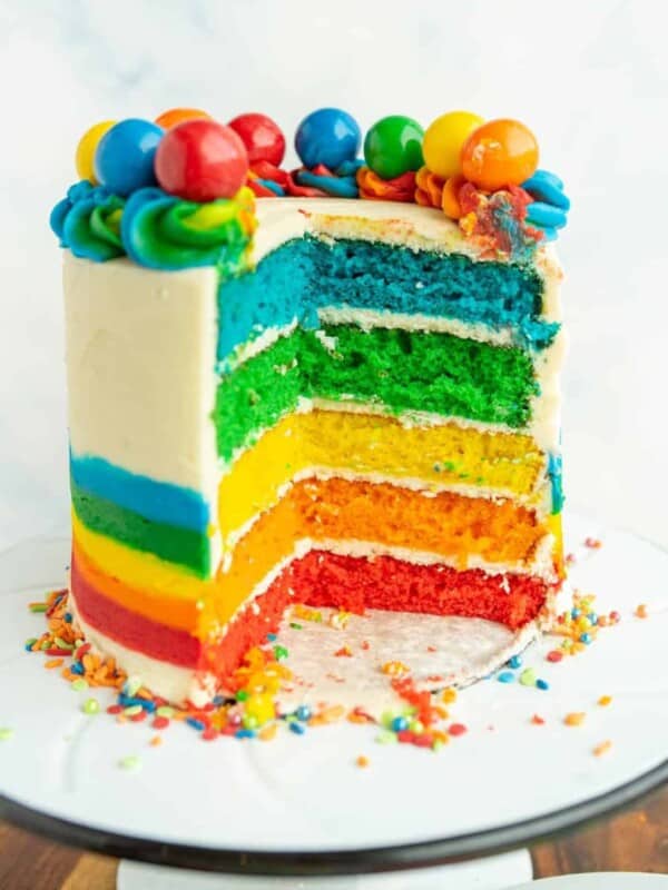 close up of the round rainbow layer cake on a white stand - the outer icing is decorated with the bottom half as a rainbow and top 3/4 white icing and along the top of the cake is rainbow piped dollops with different colorful gumboils on top