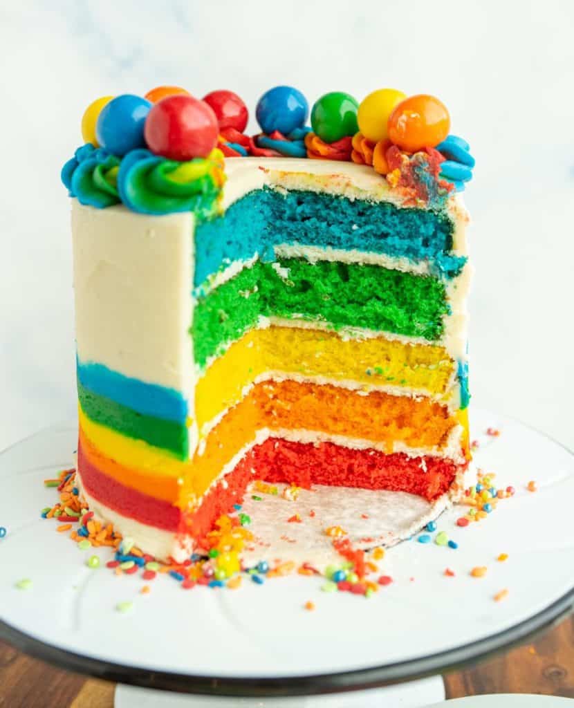 close up of the round rainbow layer cake on a white stand - the outer icing is decorated with the bottom half as a rainbow and top ¾ white icing and along the top of the cake is rainbow piped dollops with different colorful gumboils on top