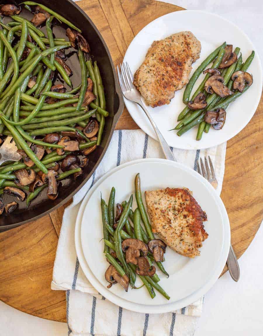 cast iron skillet with cooked long green beans and browned sliced mushrooms in the sitting to the side of a round wooden block with a towel - two plates and two forks have the vegetables on the side and a cooked pork chop lightly seasoned next to it