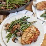 cast iron skillet with cooked long green beans and browned sliced mushrooms in the background in the foreground is a white plate with the vegetables on the side and a cooked pork chop lightly seasoned next to it
