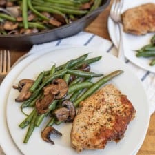 cast iron skillet with cooked long green beans and browned sliced mushrooms in the background in the foreground is a white plate with the vegetables on the side and a cooked pork chop lightly seasoned next to it