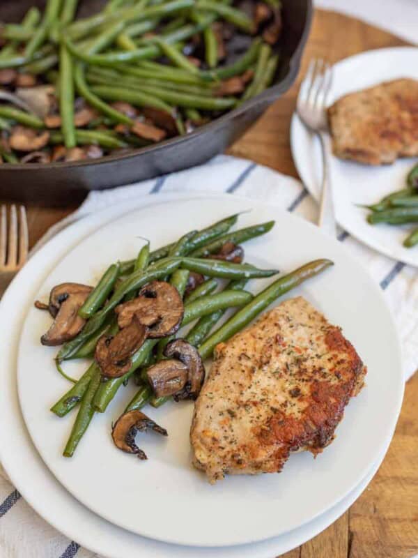 cast iron skillet with cooked long green beans and browned sliced mushrooms in the background in the foreground is a white plate with the vegetables on the side and a cooked pork chop lightly seasoned next to it