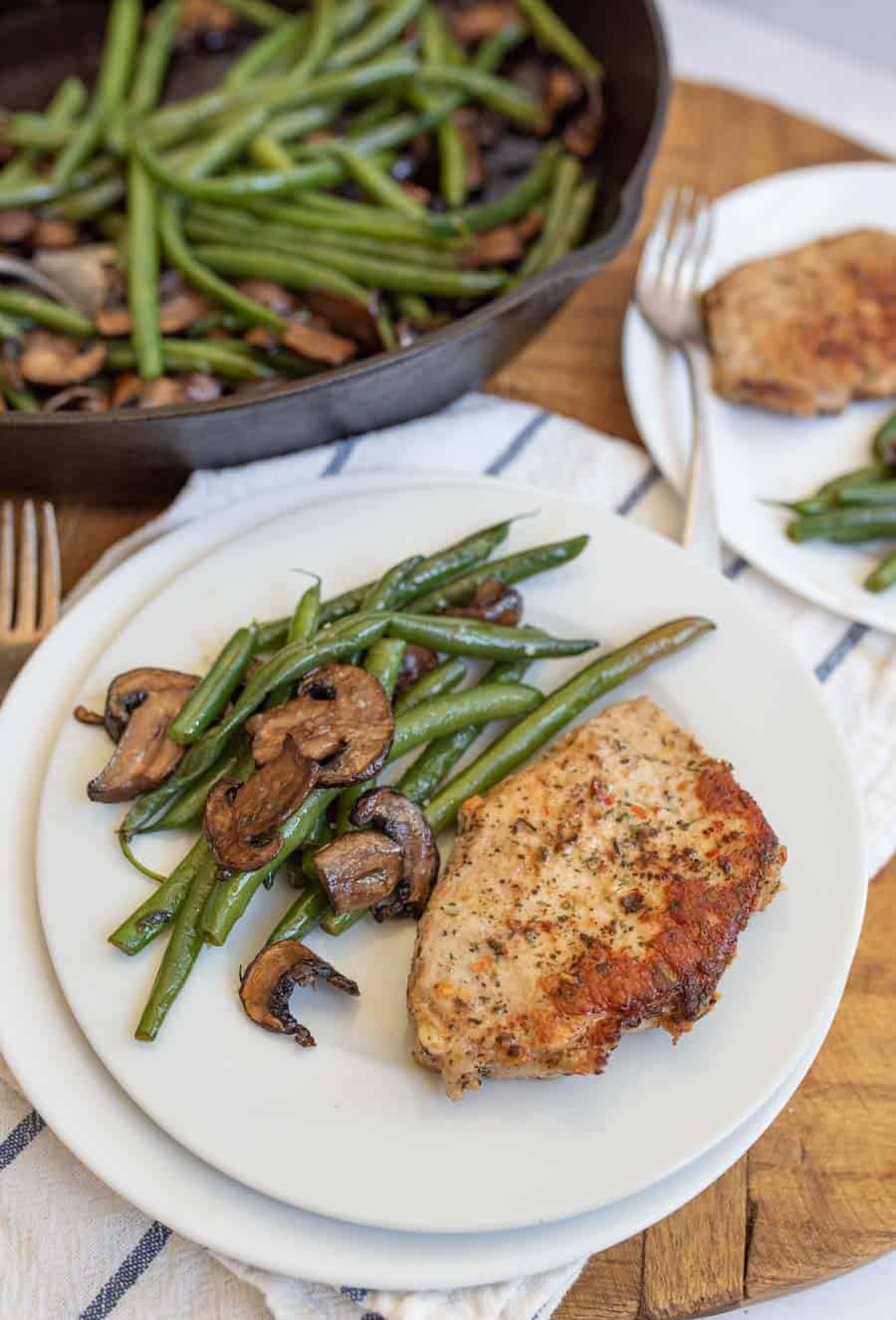 cast iron skillet with cooked long green beans and browned sliced mushrooms in the background in the foreground is a white plate with the vegetables on the side and a cooked pork chop lightly seasoned next to it