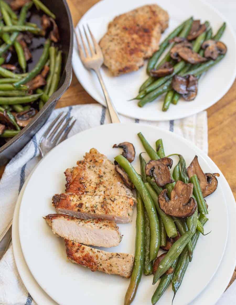 closer view of the pork dinner on a white plate a second dish and the edge of the cast iron pan in the background - beans look tender and the mushrooms are sliced and browned while the pock chop has crispy edges and various size seasonings it is cut to show a soft white center