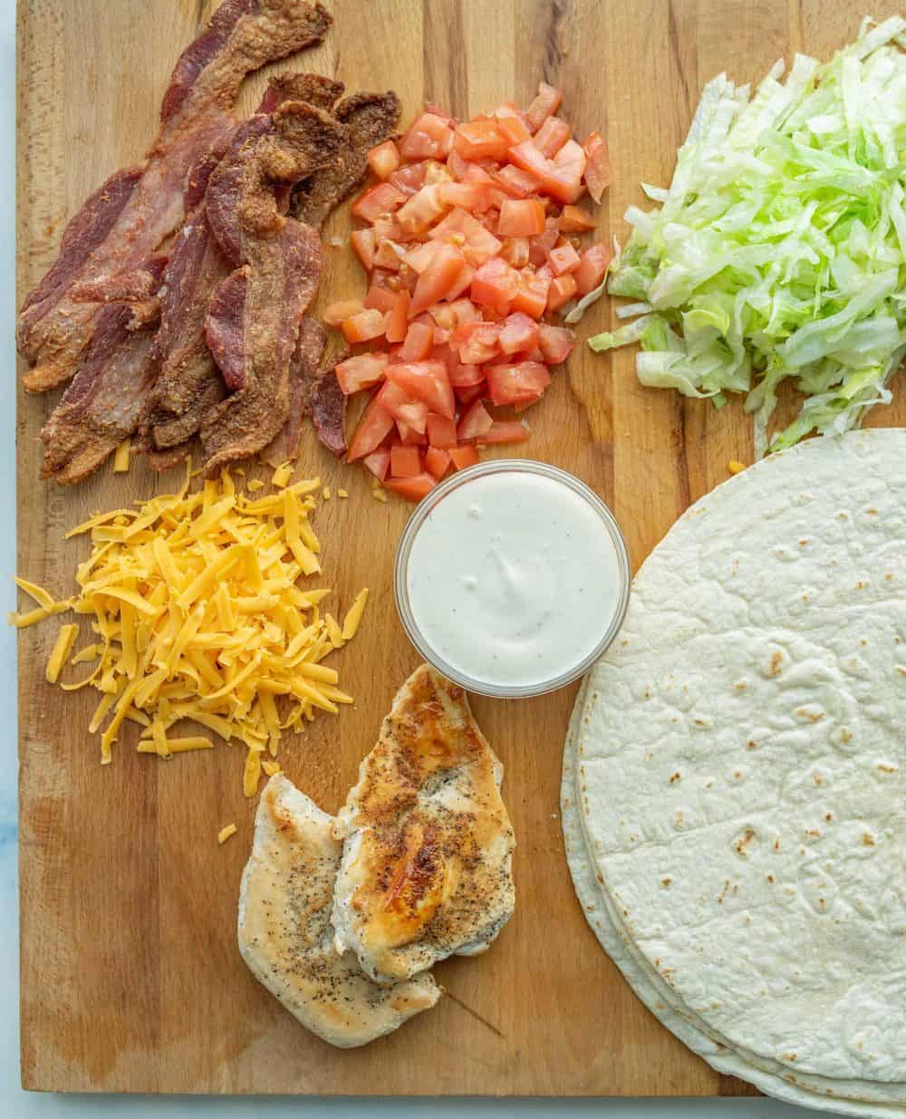 two tortilla wraps with assorted vegetables, cheese, and bacon on cutting board with piles of cheese, tomatoes, and lettuce.