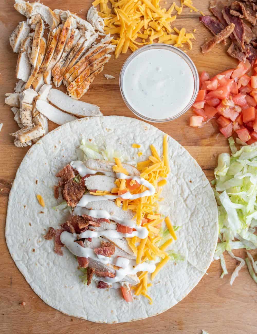 two tortilla wraps with assorted vegetables, cheese, and bacon on cutting board with piles of cheese, tomatoes, and lettuce.