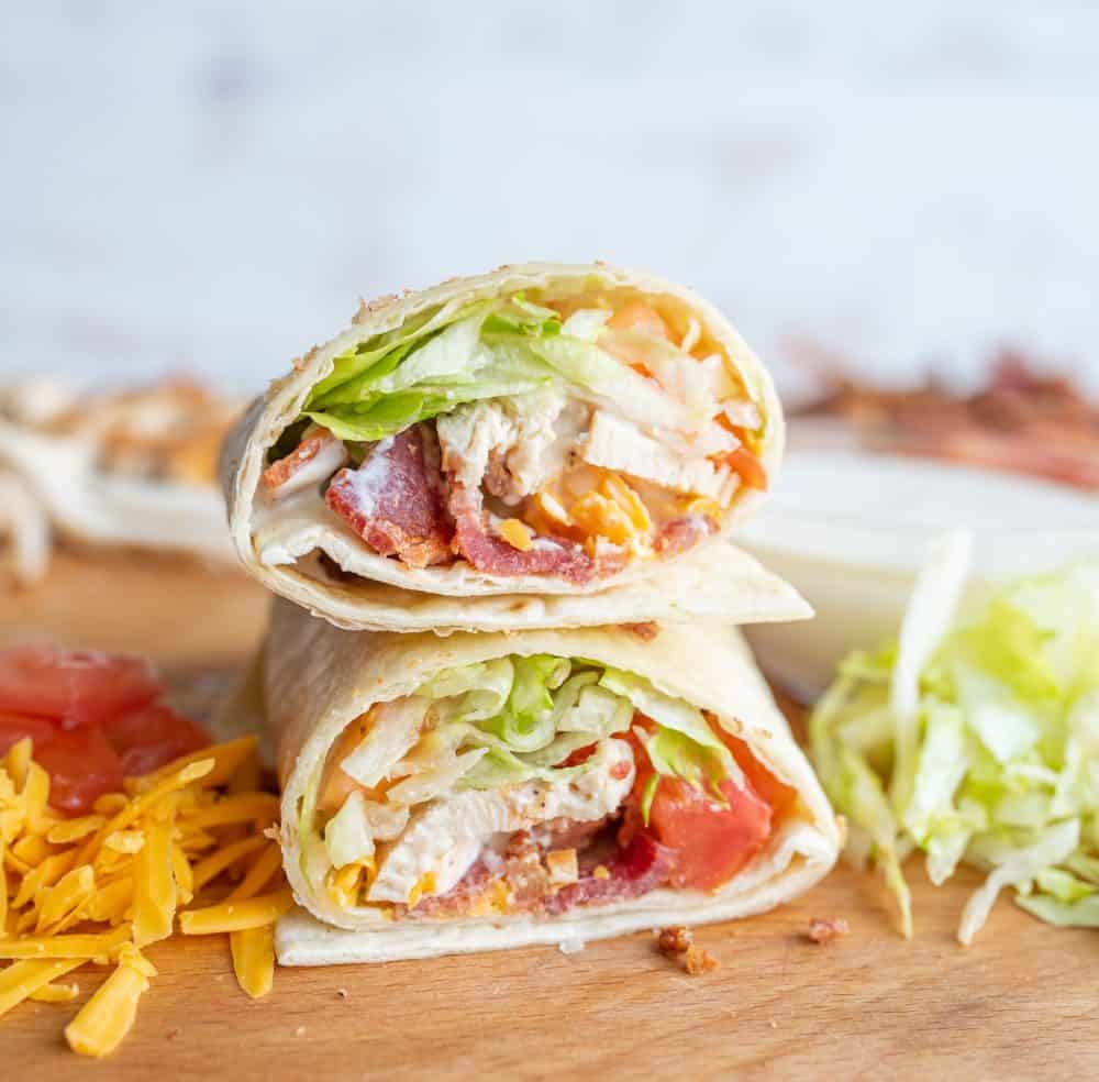 two tortilla wraps with assorted vegetables, cheese, and bacon on cutting board with piles of cheese, tomatoes, and lettuce.