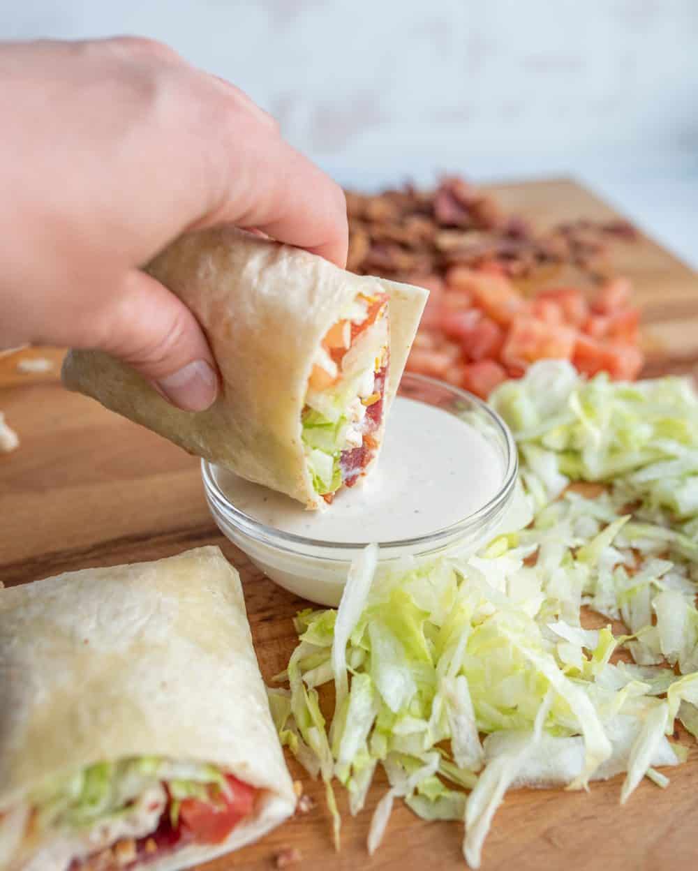 two tortilla wraps with assorted vegetables, cheese, and bacon on cutting board with piles of cheese, tomatoes, and lettuce.