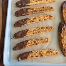 close up of biscotti on a parchment with chocolate chips and one end lightly covered in chocolate on a baking sheet pan