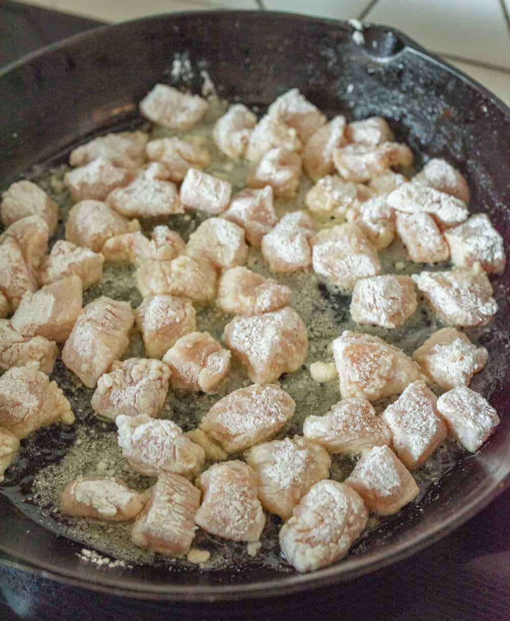 raw cubed chicken dusted in flour begening to cook in a vast iron pan.