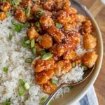 a bowl of rice with sticky sesame chicken with chives and green onion on top