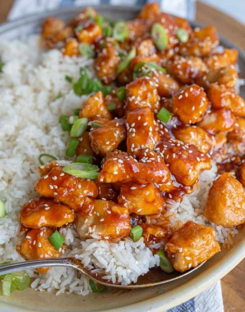 Sesame chicken in a bowl of rice, covered with sweet and tangy sauce and topped with sesame seeds and green onions.