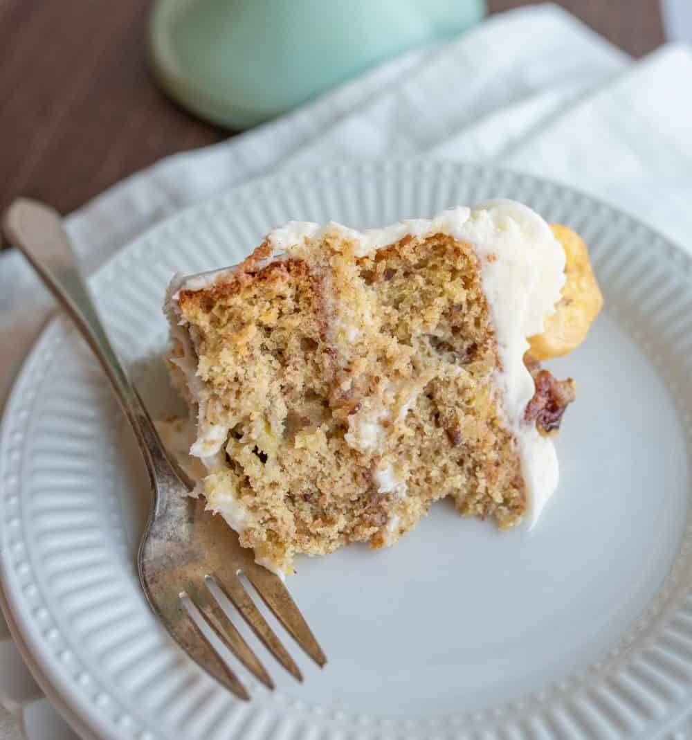 close up of a slice of humingbird cake with white frosting on a white plate and a fork.